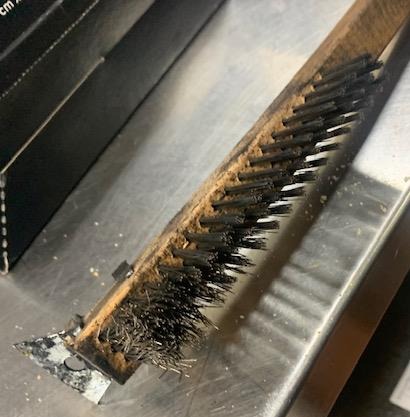 Worn consumer-grade barbecue scraper brush with bent and missing bristles on a commercial grill